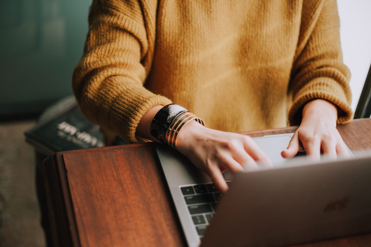 A person in a yellow seater site ad a wooden desk, typing on a computer.